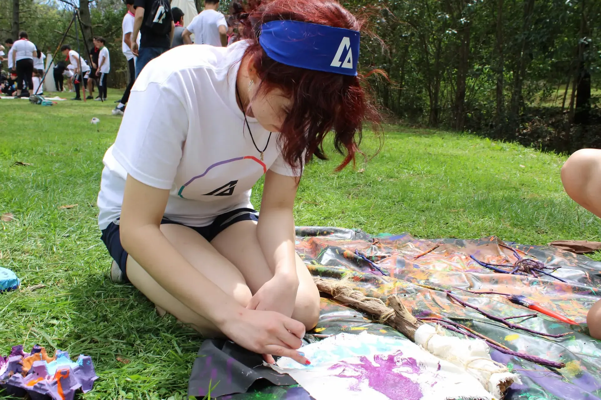 Niña con banda en la cabeza escribiendo en una hoja sentada en el pasto, durante una actividad creativa